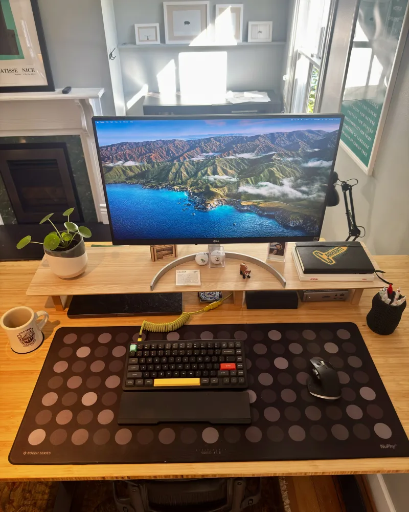 The desk by the fireplace: an LG monitor on a wooden riser, a black keyboard with a yellow space bar and coiled yellow cable, and a mouse on a polka-dot desk mat